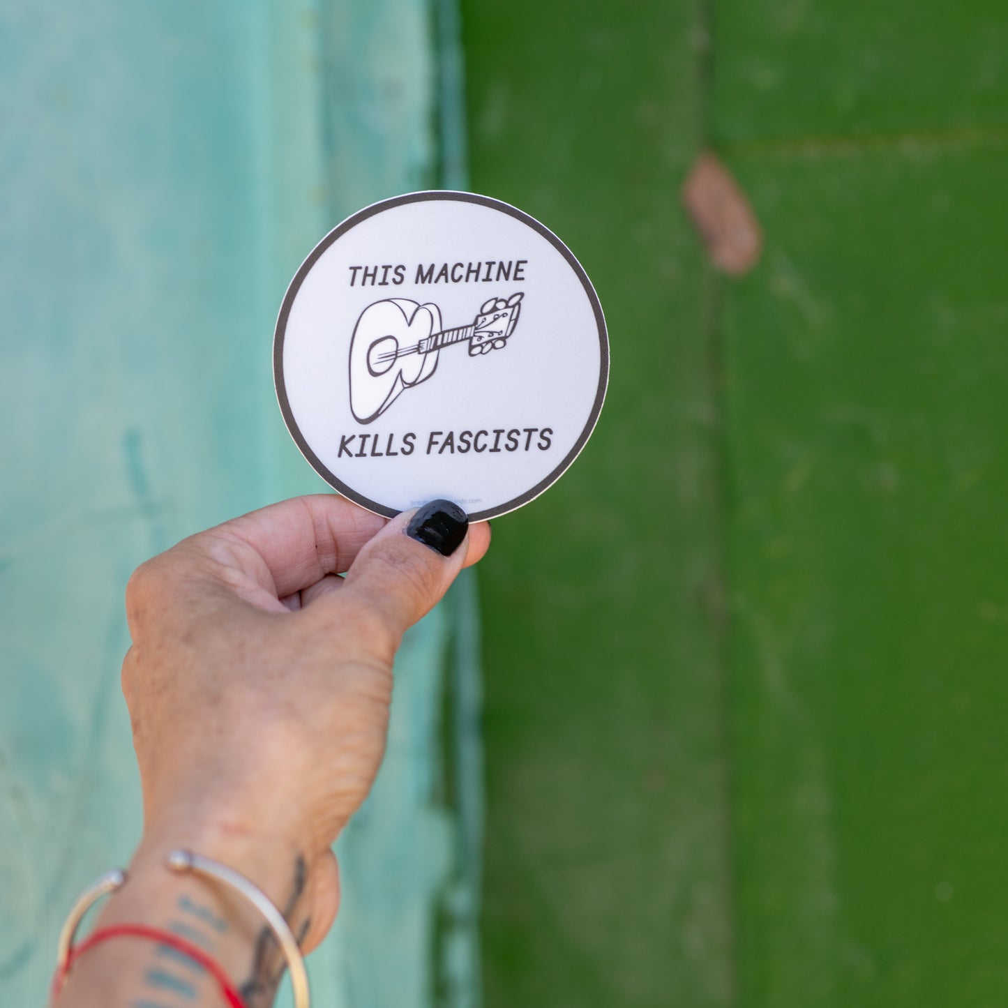 3-inch round vinyl sticker with “This Machine Kills Fascists” text — inspired by Woody Guthrie’s iconic guitar message — matte, waterproof, and UV coated. Shot against a textured background.