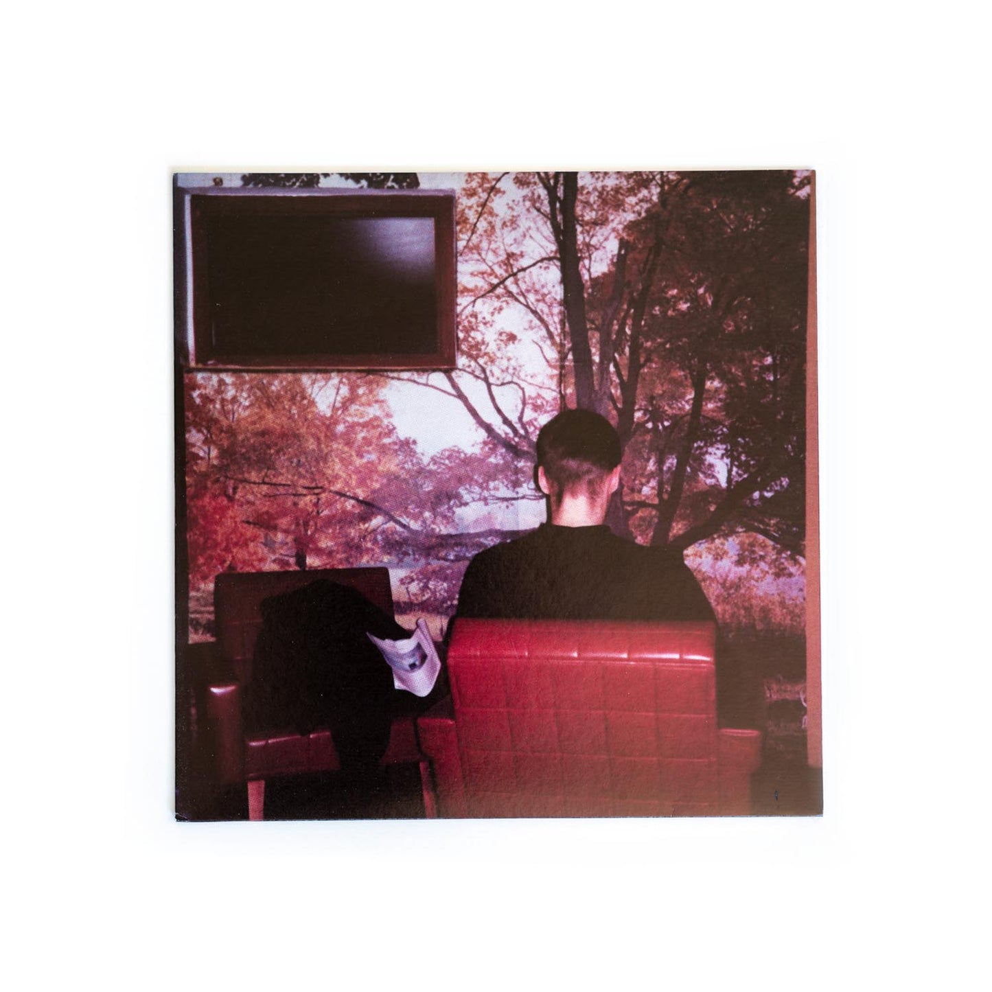 Back cover of Fugazi 7 inch Furniture. A photograph of a person sitting with their back to the camera. The wallpaper on the wall is a nature scene and a tv set hangs on the wall.